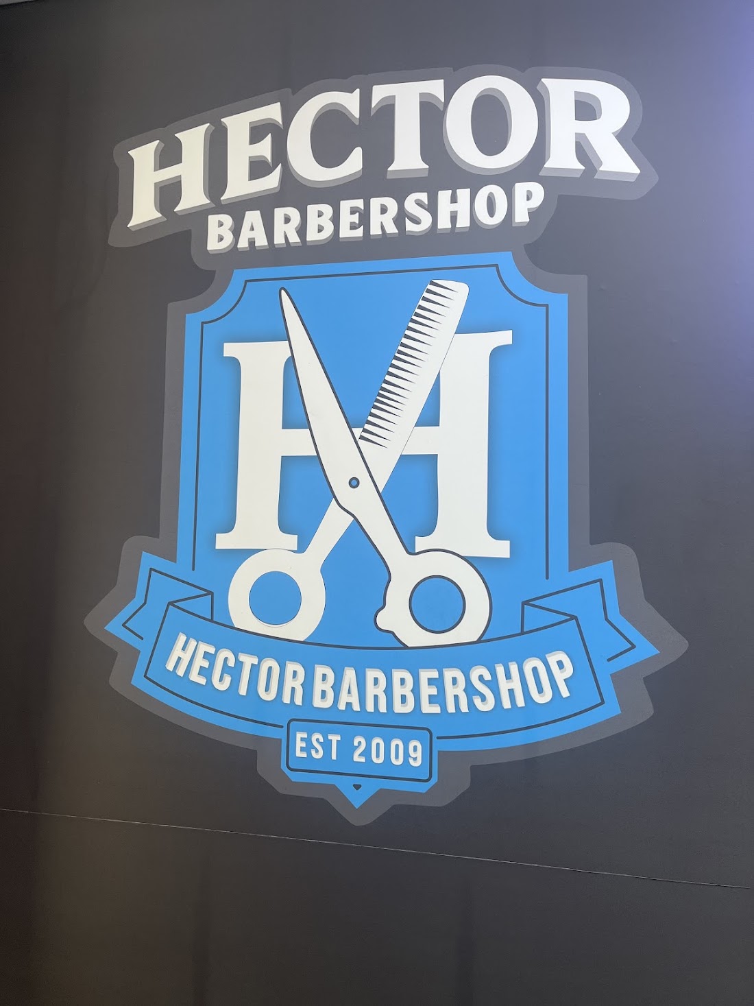 Logo de Hector Barbershop: escudo azul con tijeras y peine, establecida en 2009